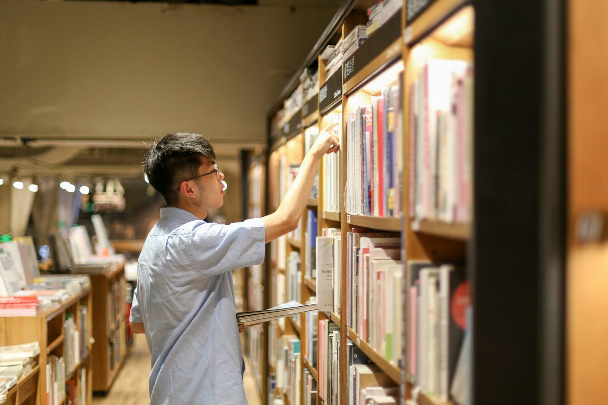 A profile shot of a young man wearing glasses and a short-sleeved light blue button-down shirt standing in a warmly lit bookstore aisle. He reaches his right hand up to select a book from a high wooden shelf while holding another book or notebook under his arm, surrounded by tall displays of neatly stacked books.