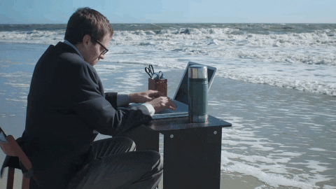 A man is typing on his laptop, seated by the beach as waves reach the shore.