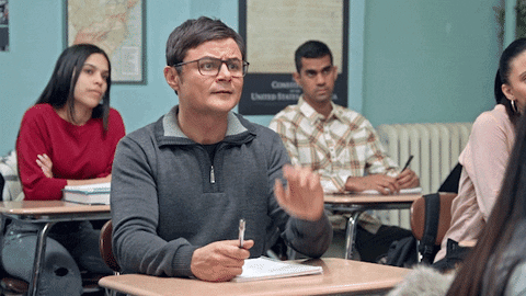 A man is raising his hand in class and saying the words, "OK. EXCUSE ME"