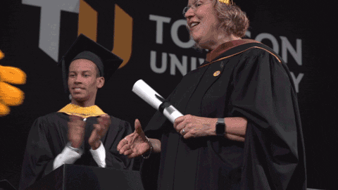 A woman in a graduation gown is standing on stage, holding a degree in one hand and shaking another person's hand with a proud smile.
