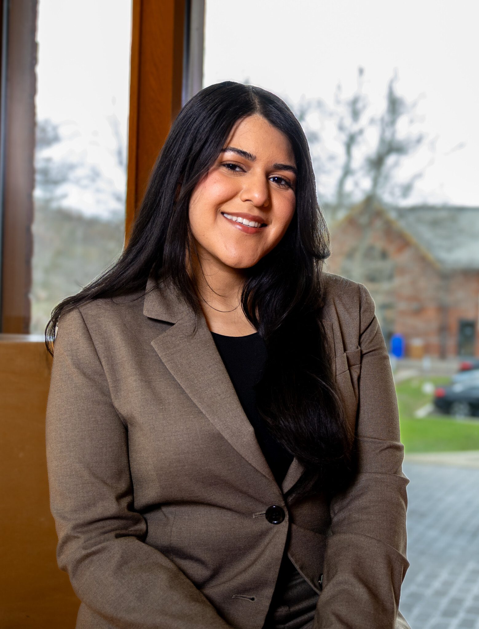 A woman with long dark hair, wearing a brown blazer and black top, sits indoors in front of large windows with blurred outdoor scenery visible in the background. She is smiling at the camera.