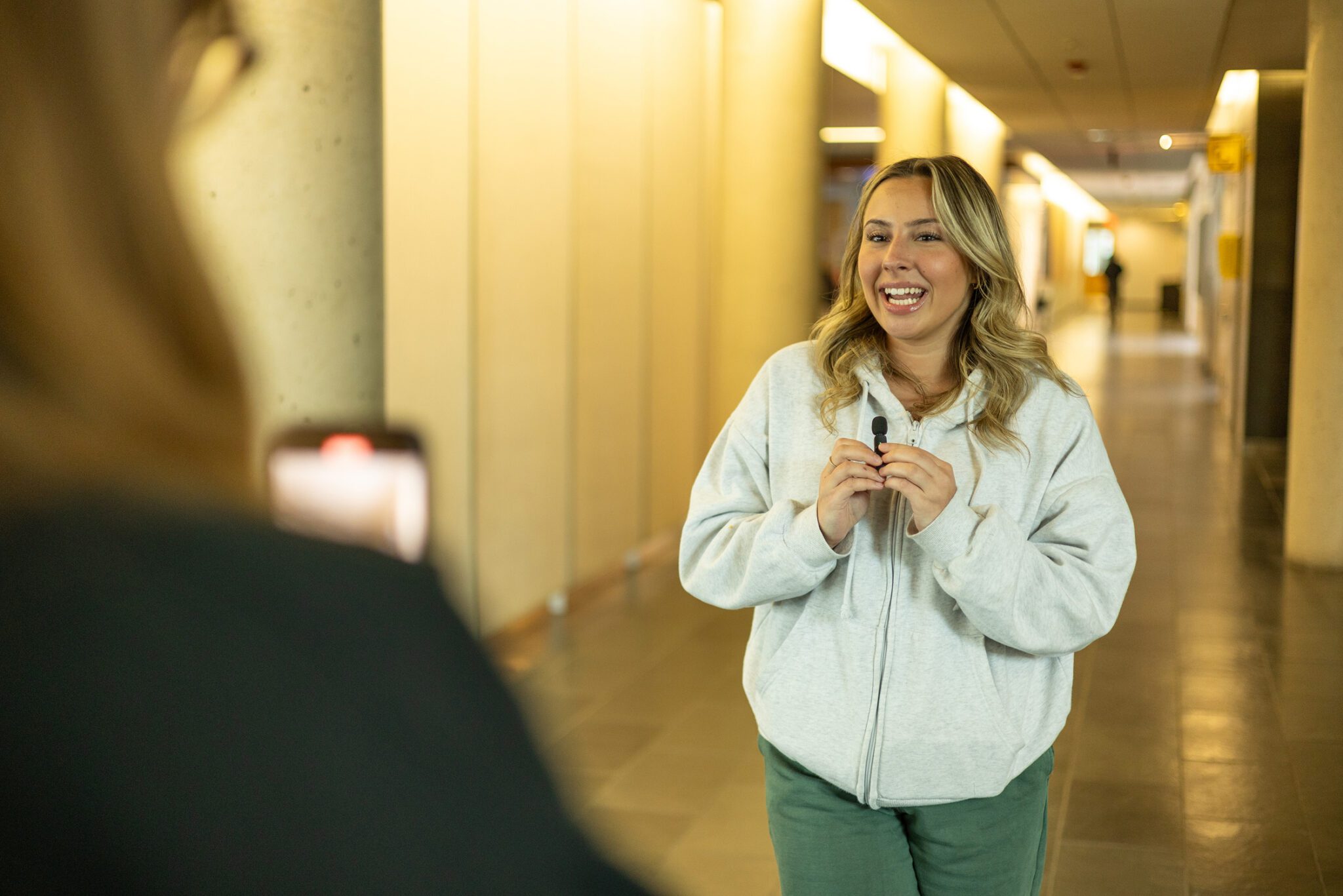 A woman in a light gray hoodie and green pants smiles and speaks into a microphone in a hallway, while another person in the foreground films her with a smartphone.