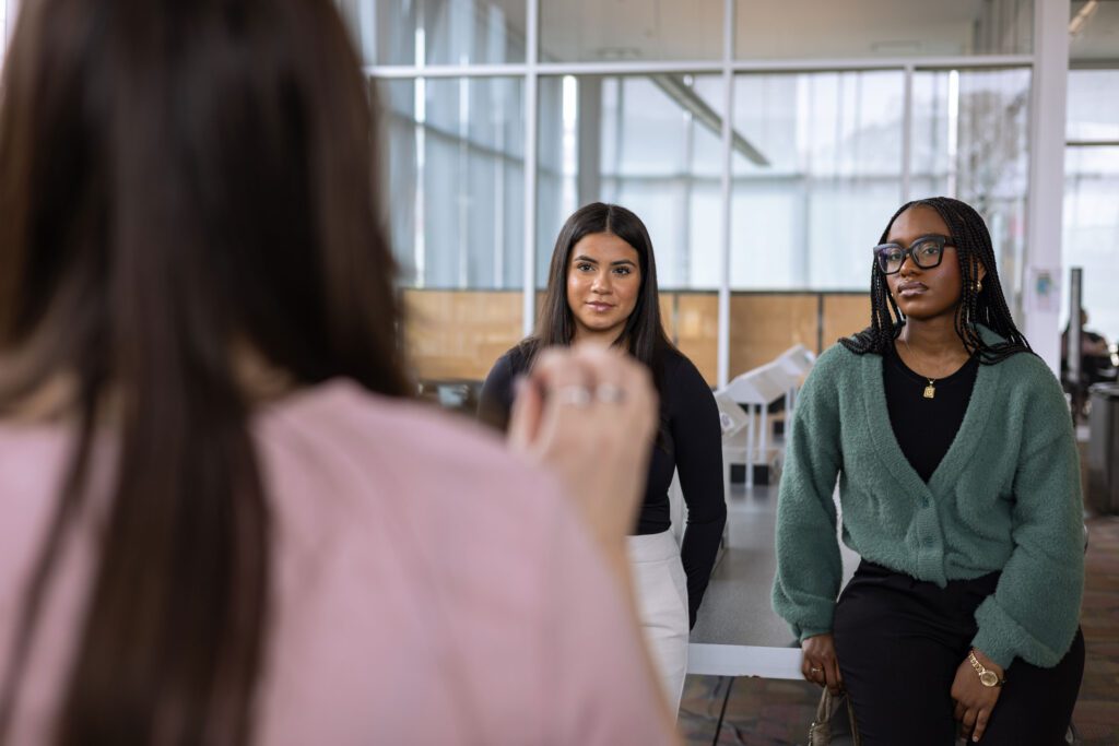 Two women stand indoors, facing a third person whose back is to the camera. One woman wears a green cardigan and glasses, the other a black top and white pants. They appear to be engaged in a conversation.