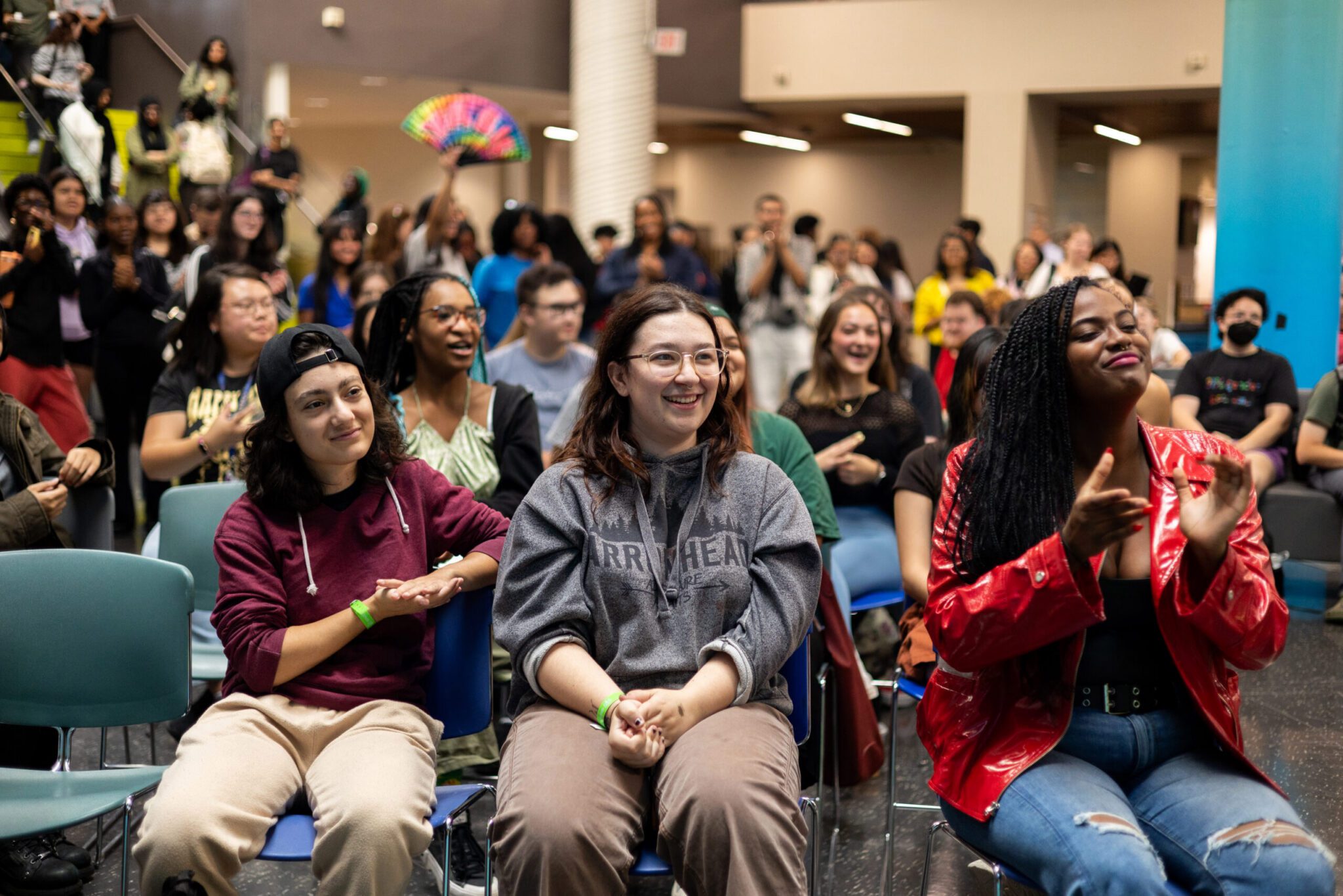 A diverse group of people sit and smile in the front row of an indoor event, while more attendees stand and interact in the background. The atmosphere is lively and cheerful.