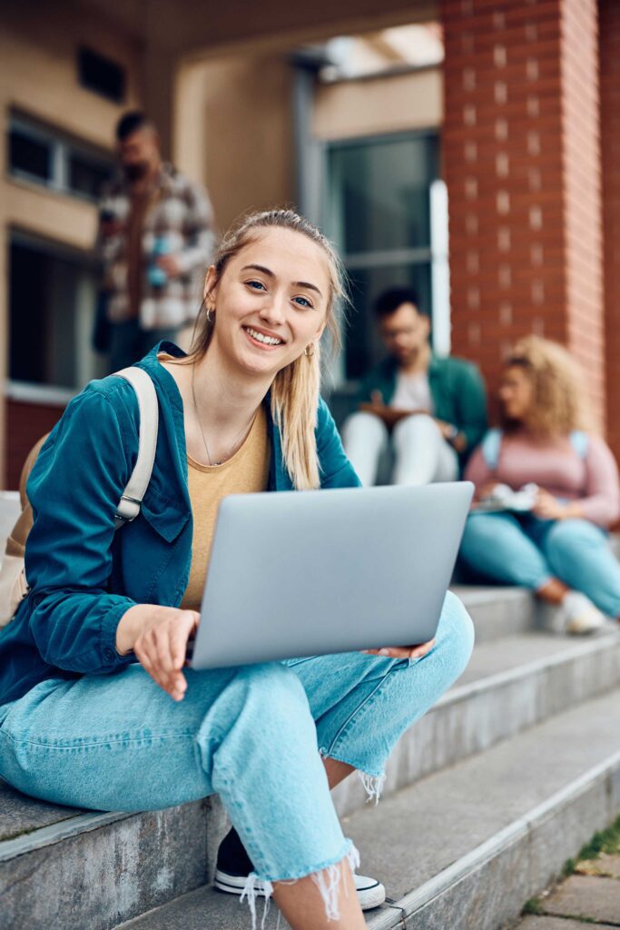 A smiling young woman with a laptop sits on outdoor steps, looking at the camera. She wears casual clothes and a backpack. Three other students are in the background, sitting and chatting near a brick building.