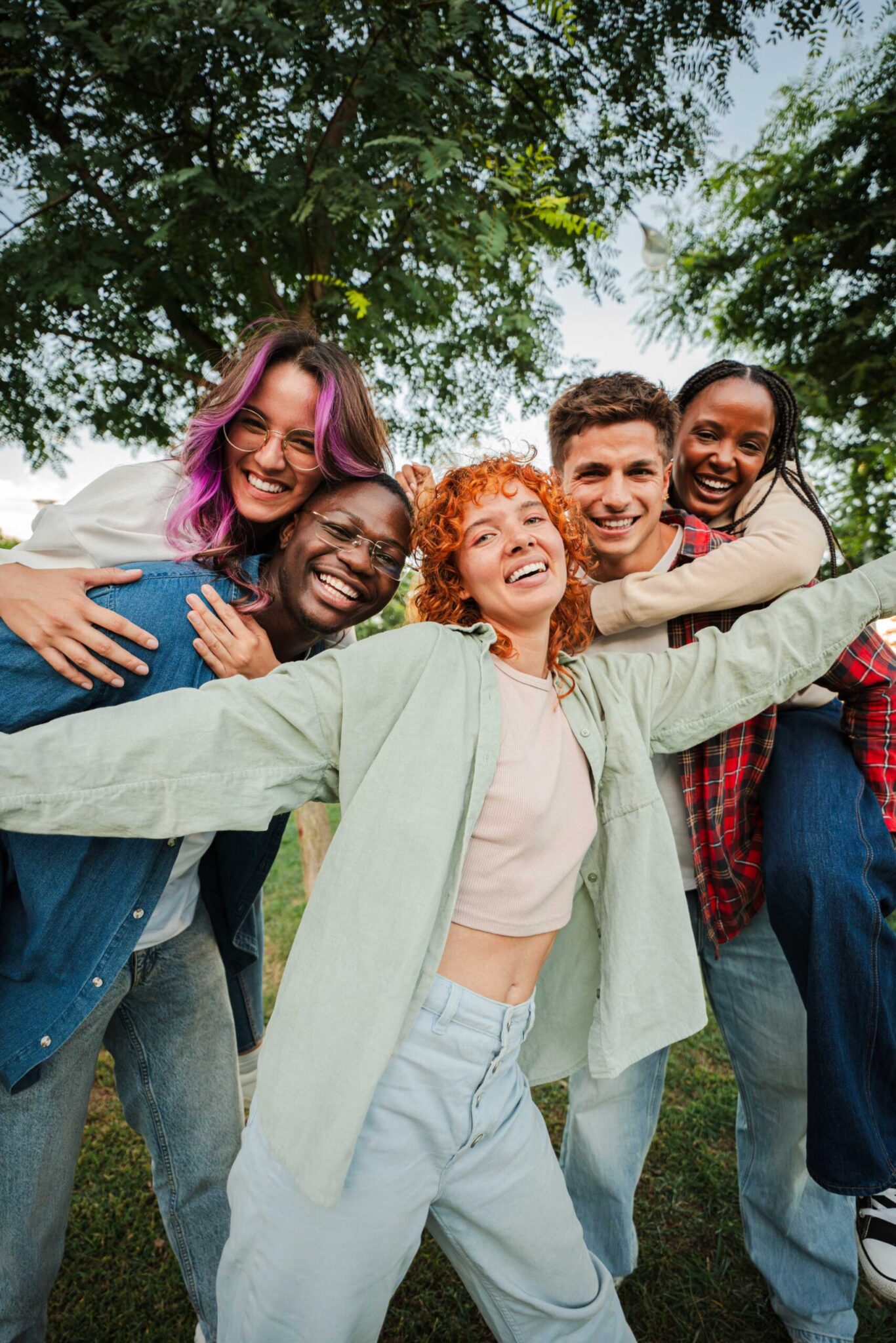 Five young adults stand close together outdoors, smiling and hugging each other. They appear happy and cheerful, with trees and greenery in the background.