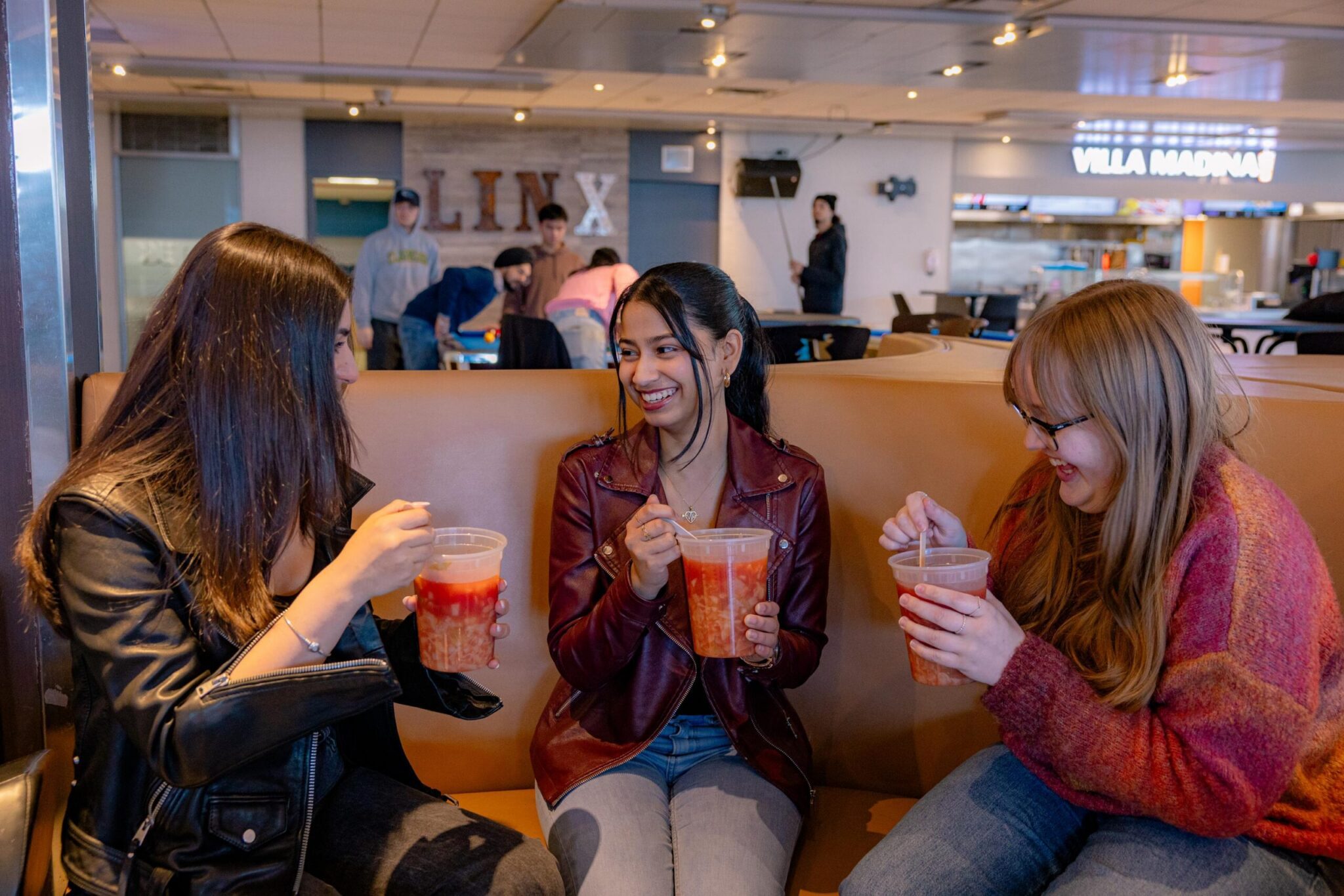 Three young women sit on a bench in a casual restaurant, smiling and talking while holding large cups of drinks with fruit pieces, enjoying each others company.