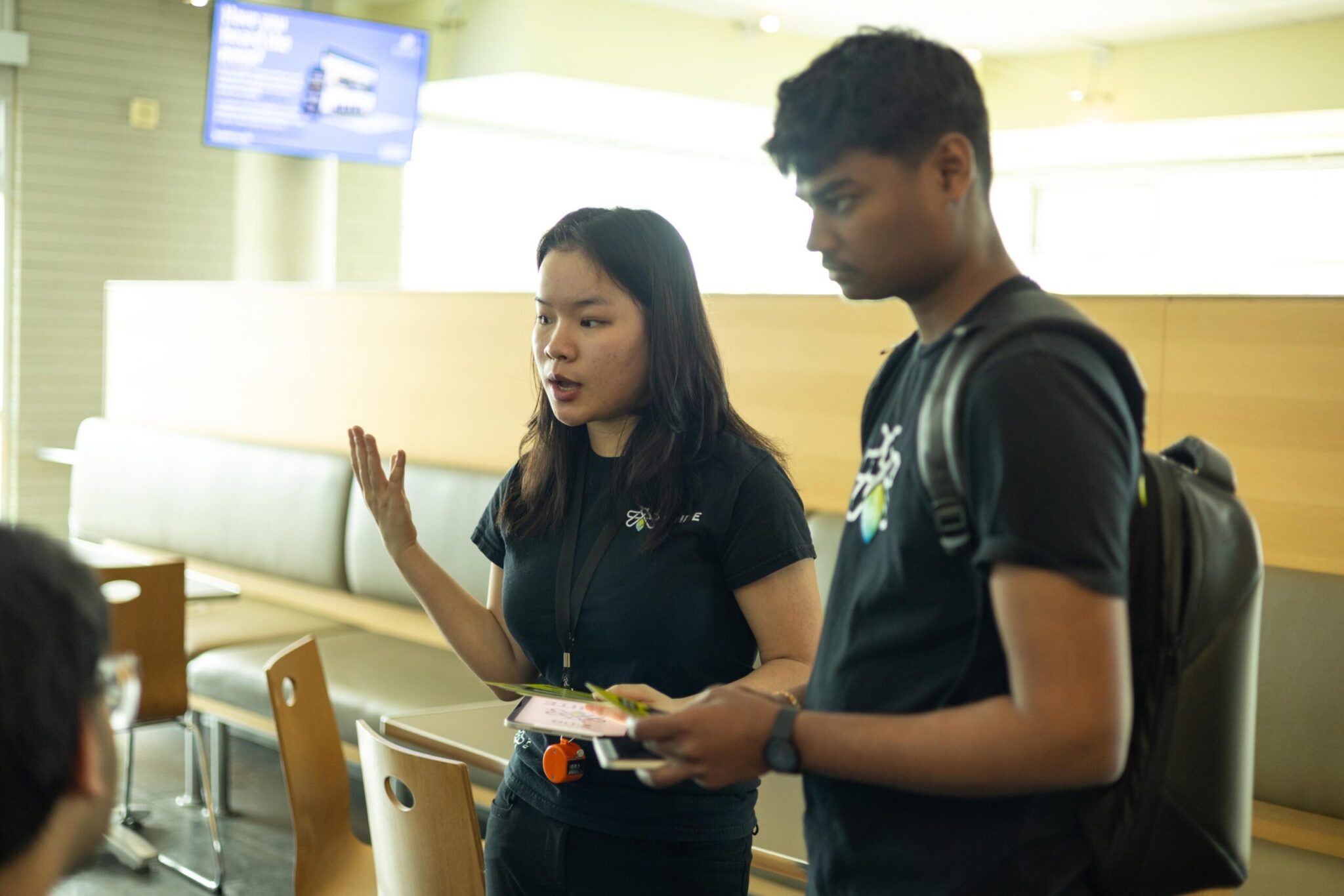 Two young adults stand indoors; one gestures while speaking, holding papers, and the other listens attentively while holding a tablet. Both wear black shirts and appear focused in a casual, modern setting with tables and chairs.