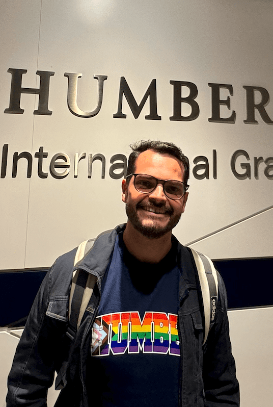 A man, wearing a HUMBER Pride t-shirt, is smiling. 