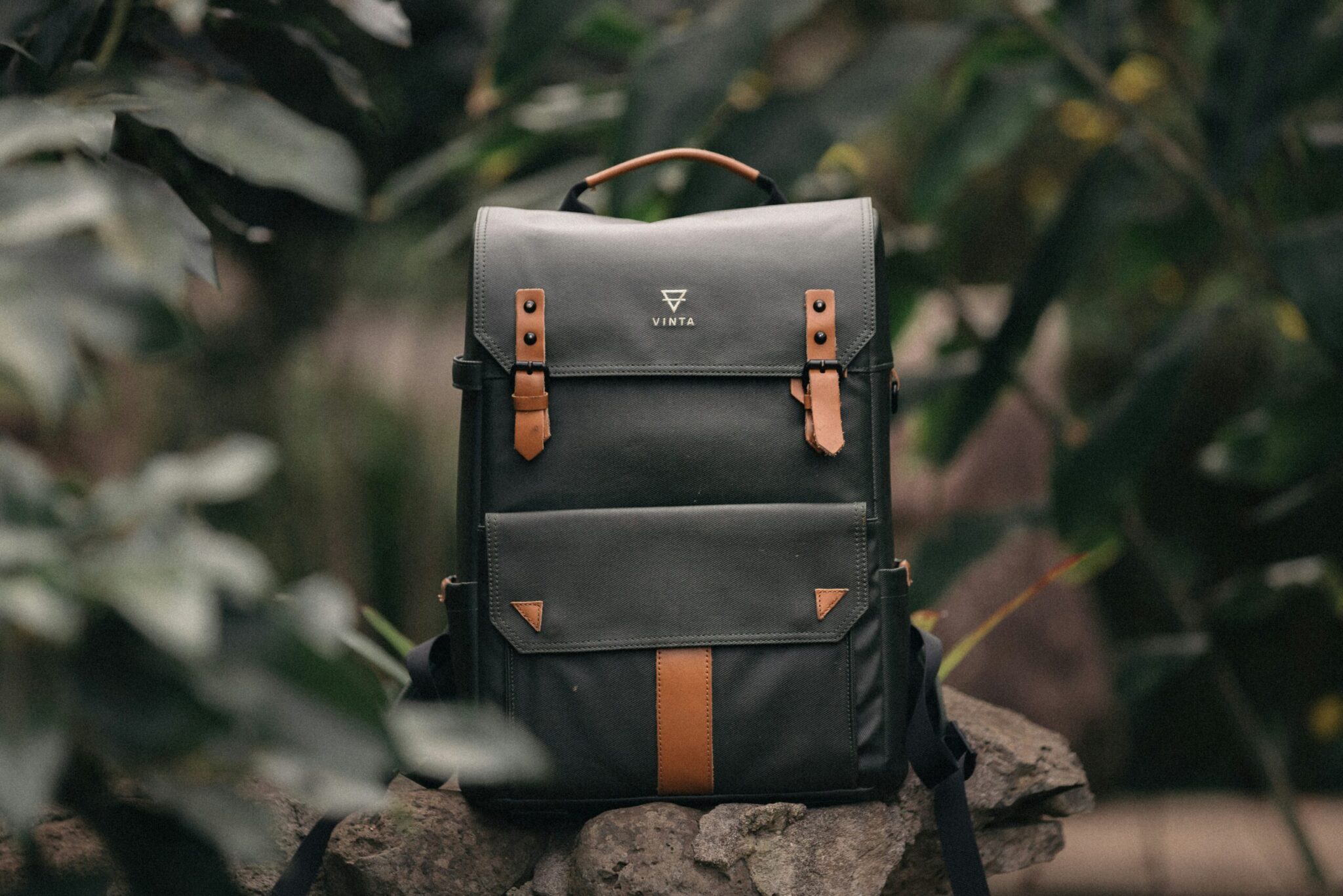 A stylish, dark charcoal grey Vinta backpack sitting upright on a rough grey rock. The backpack features light brown leather straps with black buckles on the top flap, a matching brown leather top handle, and a vertical brown leather stripe on the bottom pocket with small triangular leather details. The word "VINTA" and a triangular geometric logo are printed in gold on the upper center. The background and foreground are filled with lush, out-of-focus green foliage, creating a soft, natural outdoor setting.