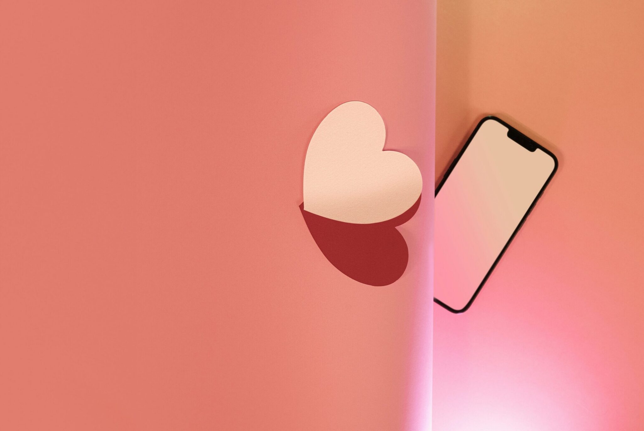 A minimalist flat lay on a split-color background of soft pink and warm peach. On the left pink section, a light pink paper heart cutout is partially lifted, casting a distinct dark heart-shaped shadow below it. On the right peach section, a smartphone with a blank white screen lies at an angle, peeking out from behind a vertical paper fold.
