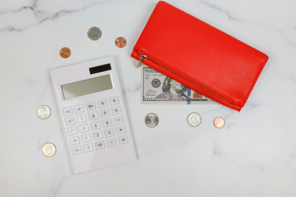 a large red wallet on top of a one hundred dollar american bill. A calculator beside it with smaller change scatter around the marble table.