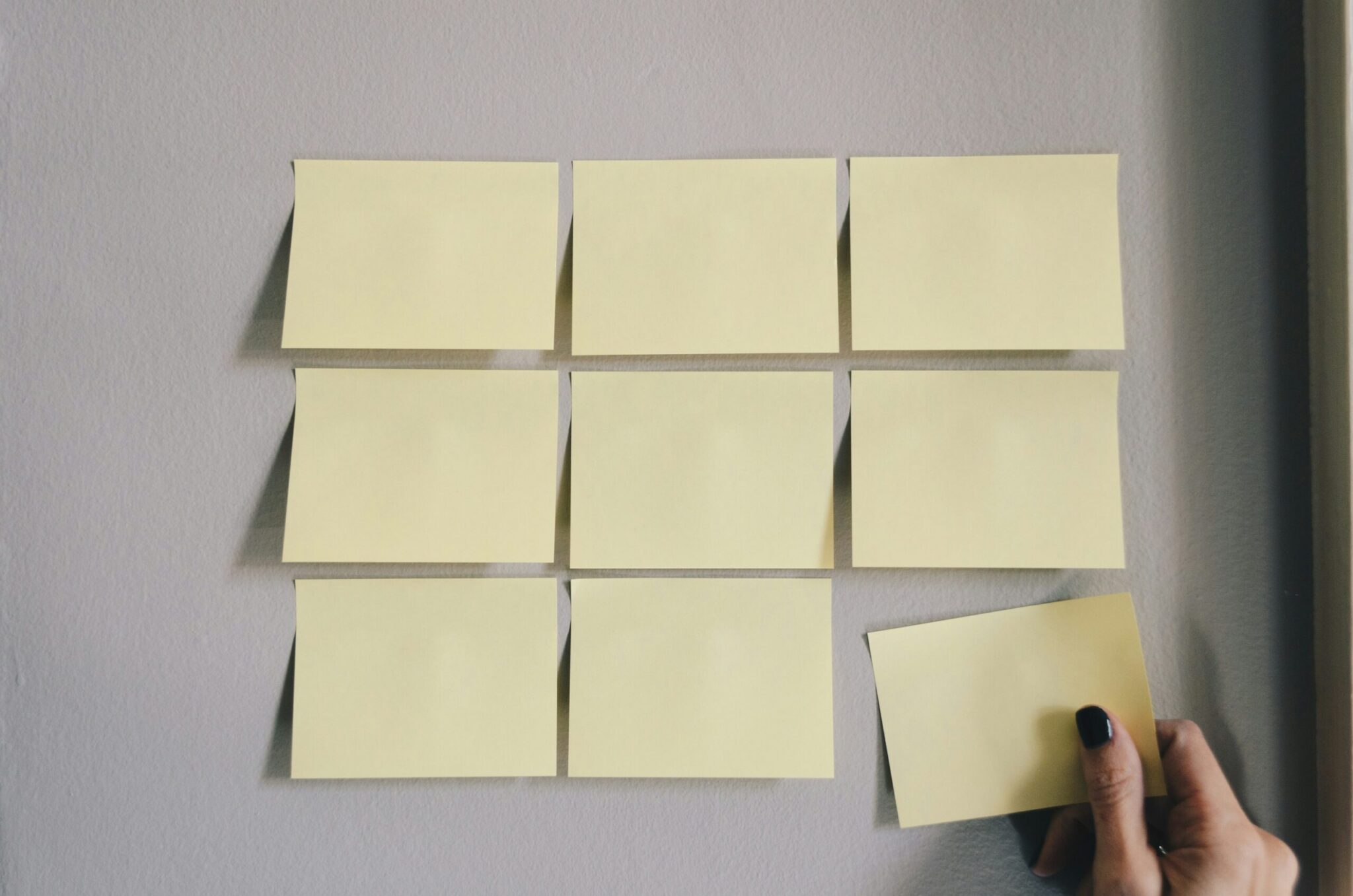 A close-up, eye-level photograph of blank pale-yellow rectangular sticky notes arranged on a textured light-grey wall. Eight of the notes are neatly aligned to form a partial $3 \times 3$ grid. In the bottom-right corner, a hand with fingernails painted with black polish holds a ninth sticky note, preparing to place it and complete the grid. Soft, natural lighting from the side casts subtle shadows under the left edges of the sticky notes, giving them a slight three-dimensional appearance.