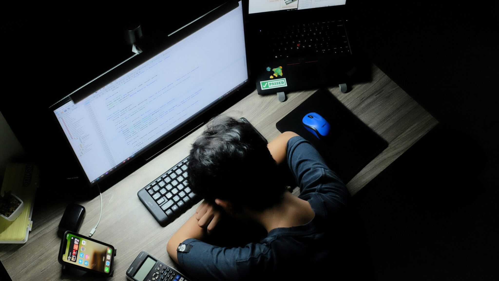 An overhead shot of a person resting their head face-down on their arms on a wooden desk in a dark room, exhausted from late-night studying or coding. They sit in front of a bright curved monitor displaying lines of programming code, an open laptop with a green "PASSED" sticker, a bright blue mouse on a mousepad, a black keyboard, a smartphone on a stand, and a scientific calculator.