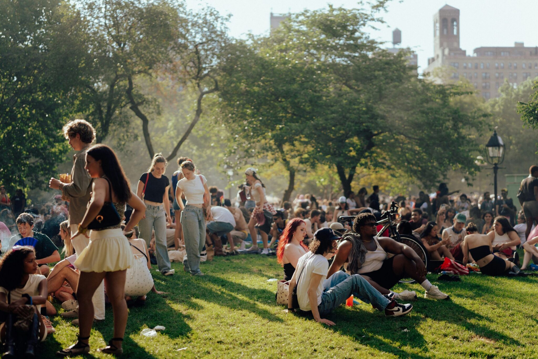 A wide shot of a bustling city park filled with people lounging, sitting, and walking across a grassy lawn on a bright, sunny day. Lush green trees frame the background with city buildings visible in the distance under soft daylight filtering through the leaves.