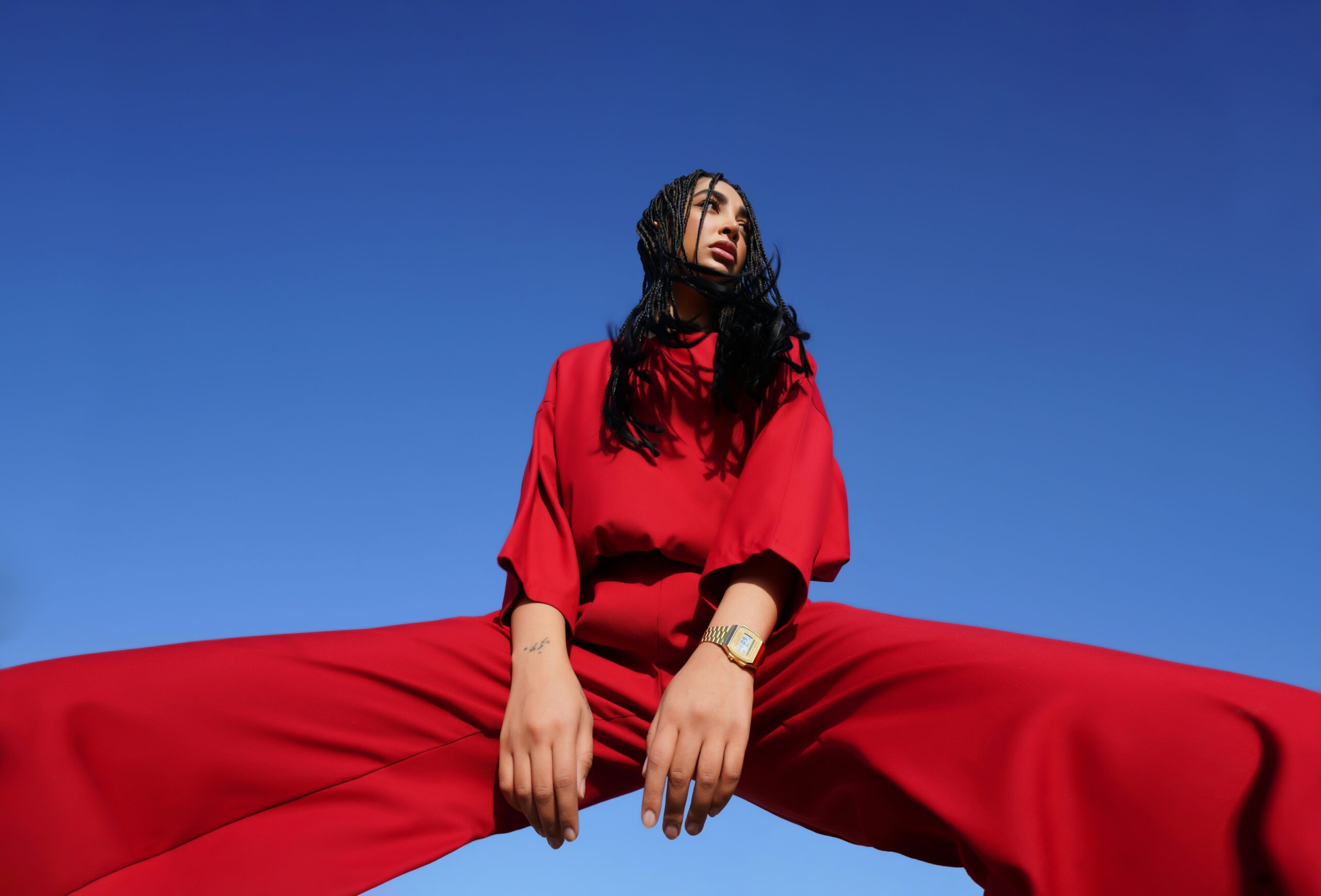 A low-angle fashion shot of a woman with braided dark hair wearing a vibrant red two-piece outfit and a gold watch. Straddling the camera with wide-spread legs against a clear blue sky, her hands rest forward as she looks thoughtfully toward the upper right.