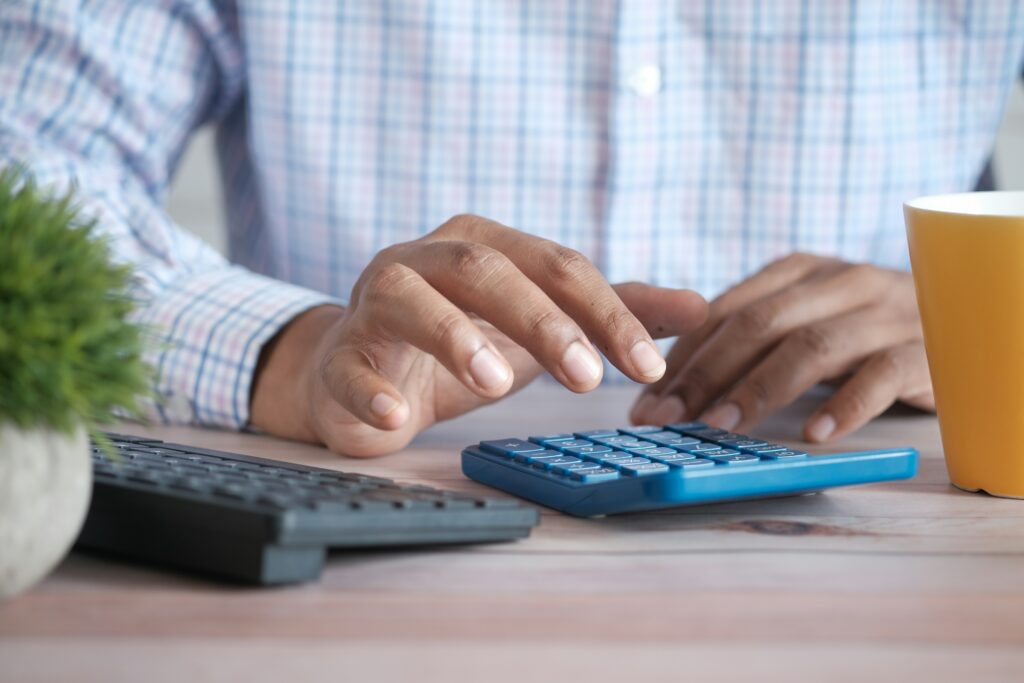 A person in a plaid shirt uses a blue calculator at a wooden desk, with a keyboard, a potted plant, and a yellow cup nearby.