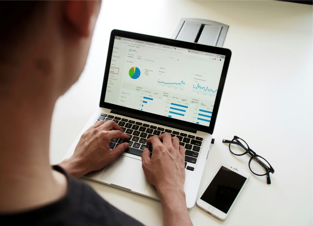 A person types on a laptop displaying charts and graphs. Next to the laptop are a pair of eyeglasses and a smartphone on a white table. The screen shows data analytics and performance metrics.