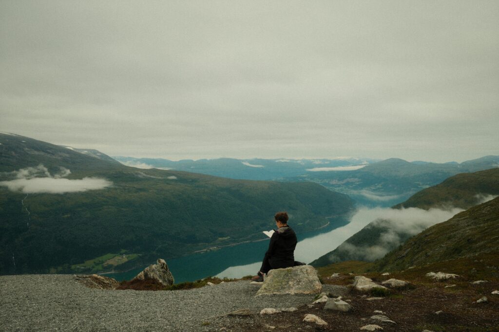 A person sitting on a rock overseeing a river and many hills while reading a book.
