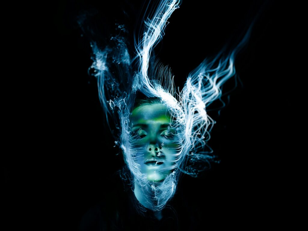 A person in an pitch black room with gleaming with neon light blue lights coming from threads wrapped around the persons head. 