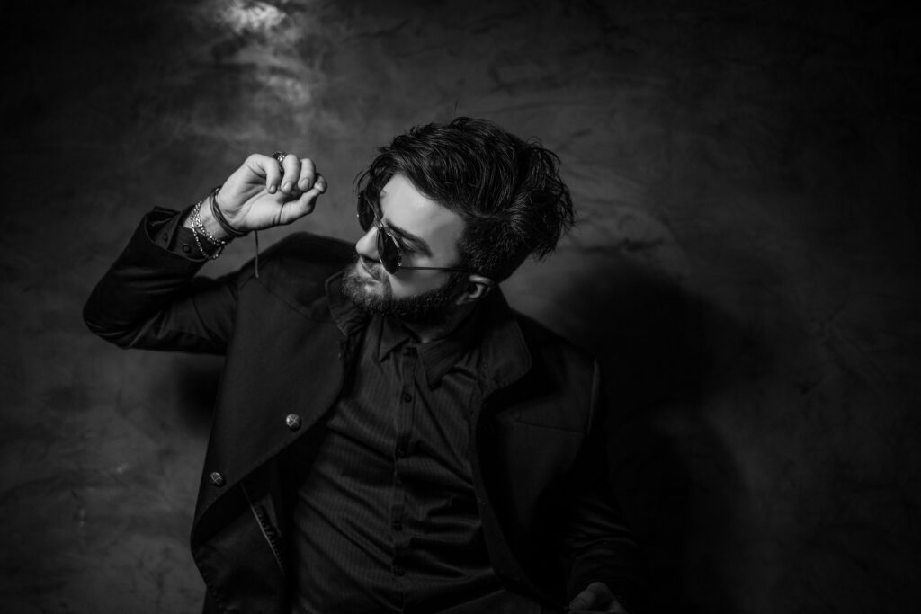 A man in black and white wearing sunglasses bracelets a dress shirt and jacket with one hand raised and his head tilted toward that hand. street