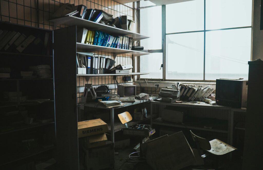A messy room filled with papers and old tech. Shelves line the walls with binders and the room filled with boxes. 