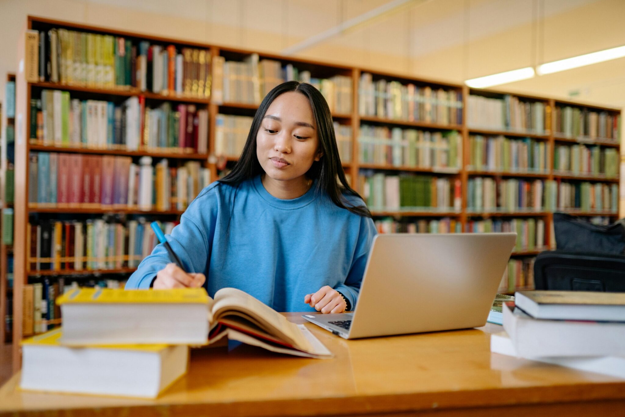 A young Asian woman with long dark hair wearing a bright blue sweatshirt sitting at a wooden table in a library. She takes notes with a blue pen in an open book while working alongside an open silver laptop, surrounded by stacks of textbooks with tall, fully stocked bookshelves spanning the background.