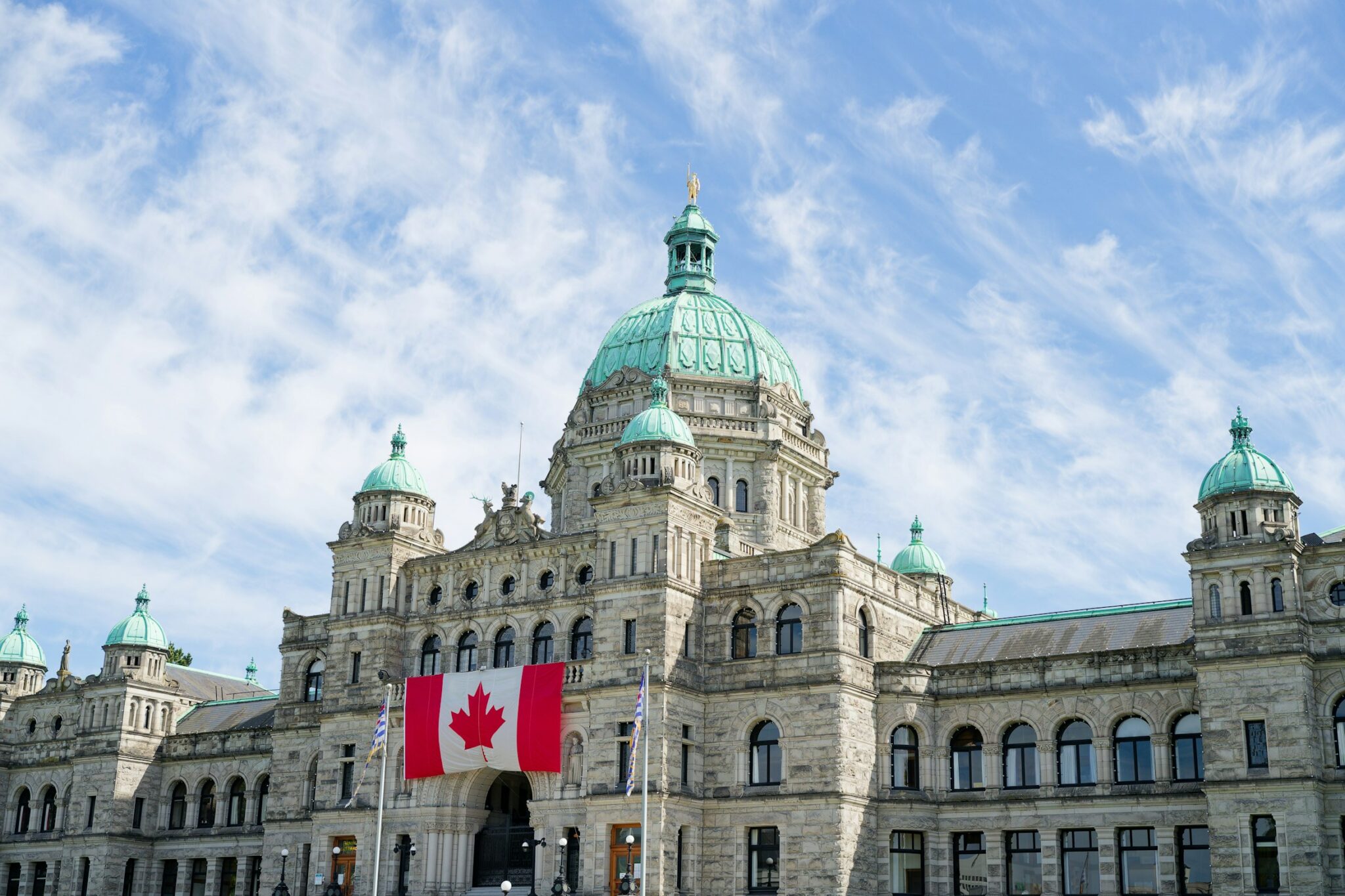A picture of parliament with a Canadian flag in the lower bottom of the picture and a blue sky with white clouds above.
