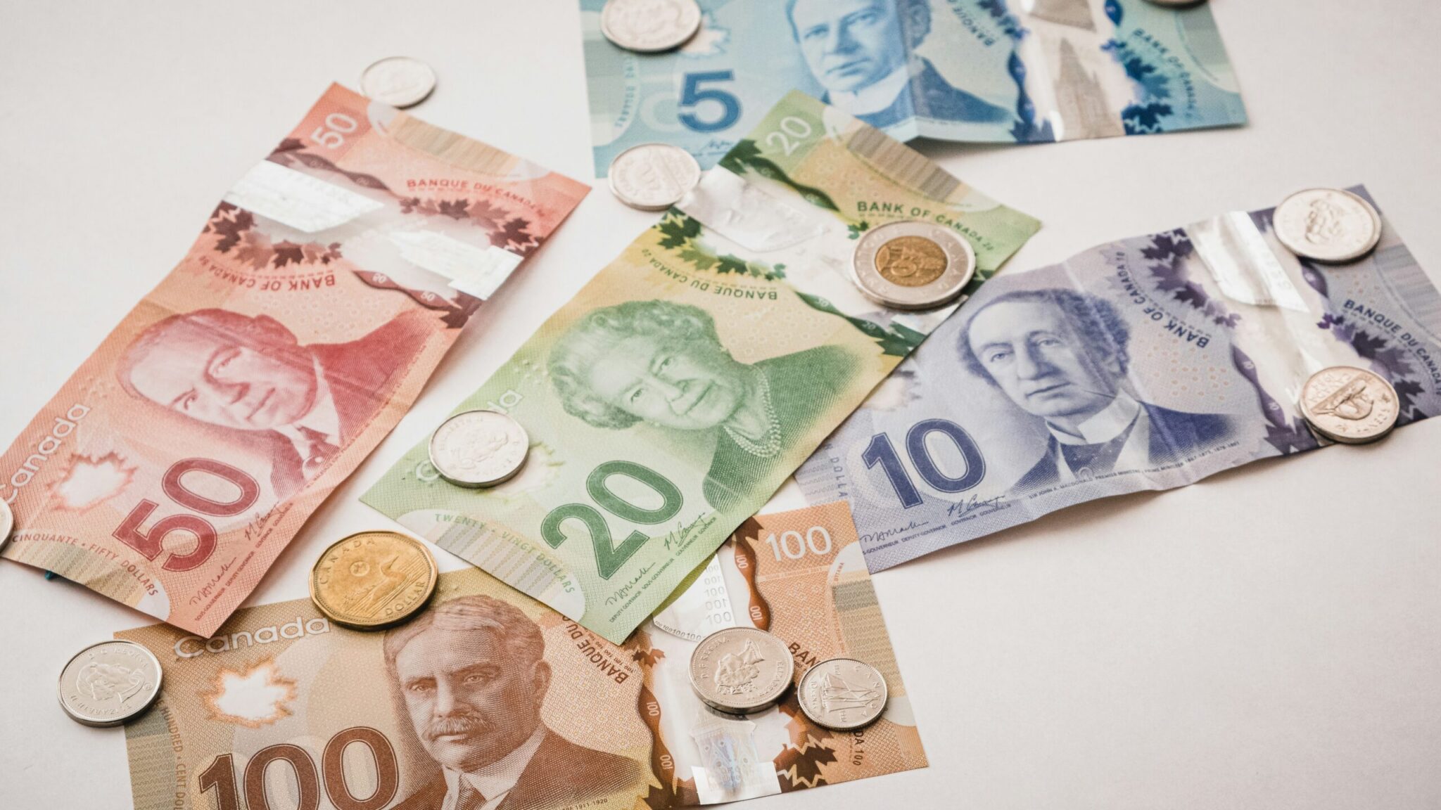 An overhead, slightly angled shot of various colorful Canadian polymer banknotes and metal coins scattered across a plain white surface. The banknotes shown include a blue five-dollar bill, a purple ten-dollar bill, a green twenty-dollar bill featuring Queen Elizabeth II, a red fifty-dollar bill, and a brown one-hundred-dollar bill. Multiple Canadian coins, including a bi-metallic gold and silver toonie ($2), a gold-colored loonie ($1), and several silver-colored quarters and dimes, are dispersed on top of and around the bills.