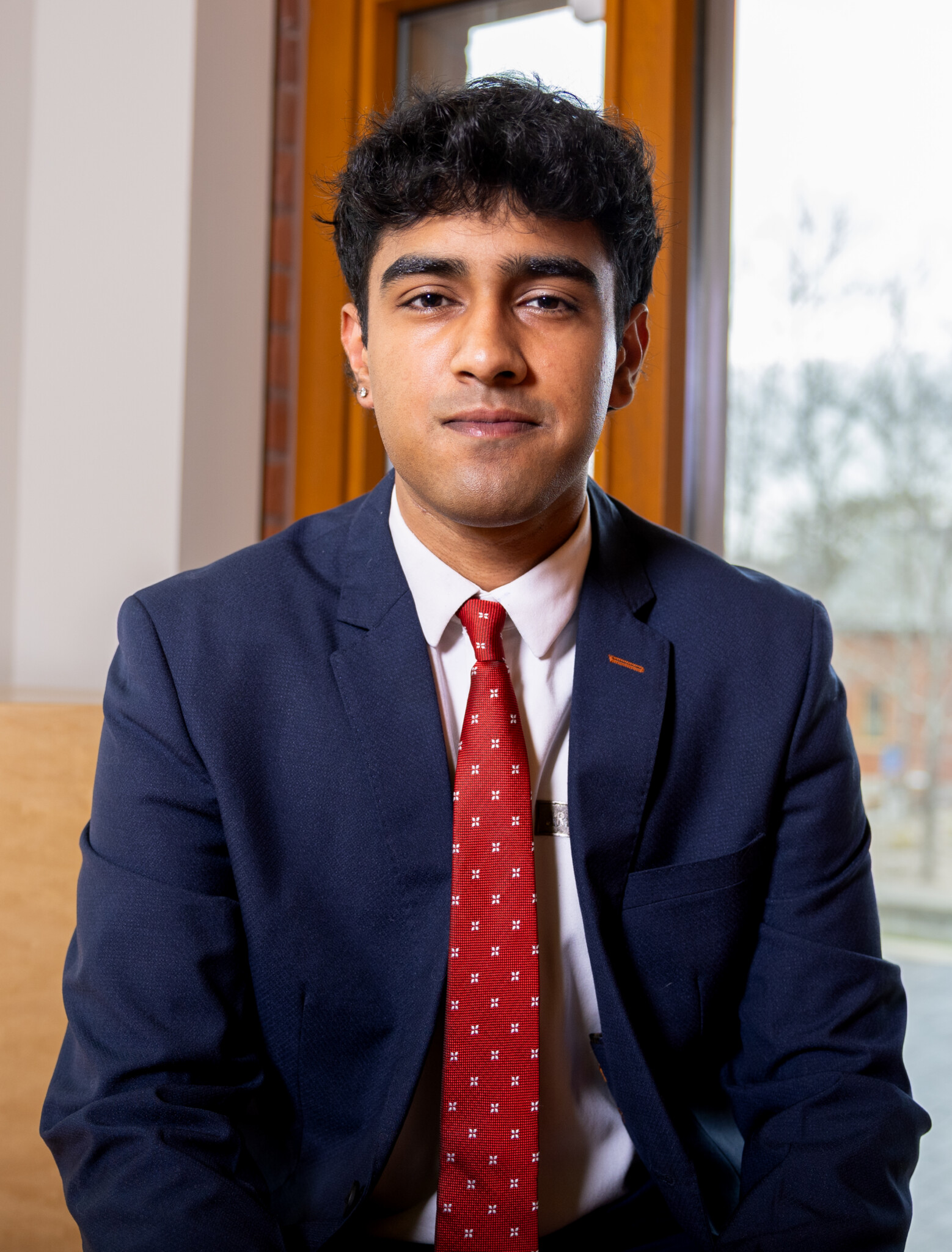 A young man with short dark hair, wearing a navy suit, white shirt, and red patterned tie, sits indoors in front of a window, looking at the camera with a neutral expression.