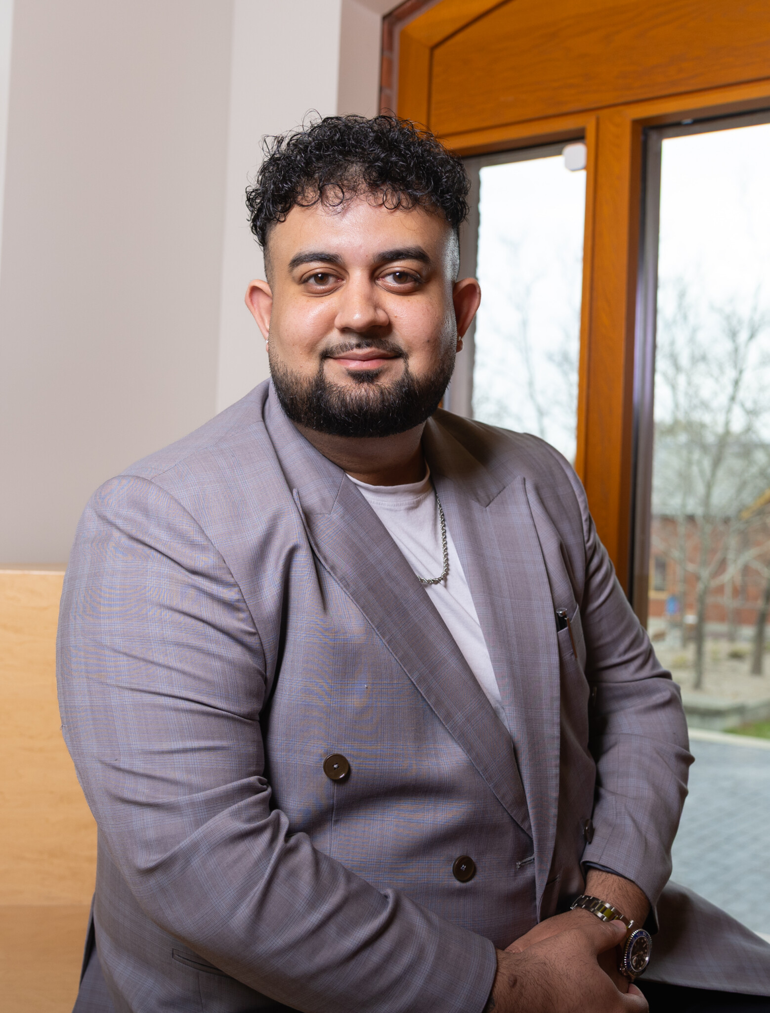 A man with short curly hair and a beard, wearing a light gray suit and white shirt, sits indoors near a wooden-framed window with trees visible outside. He is smiling and looking at the camera.