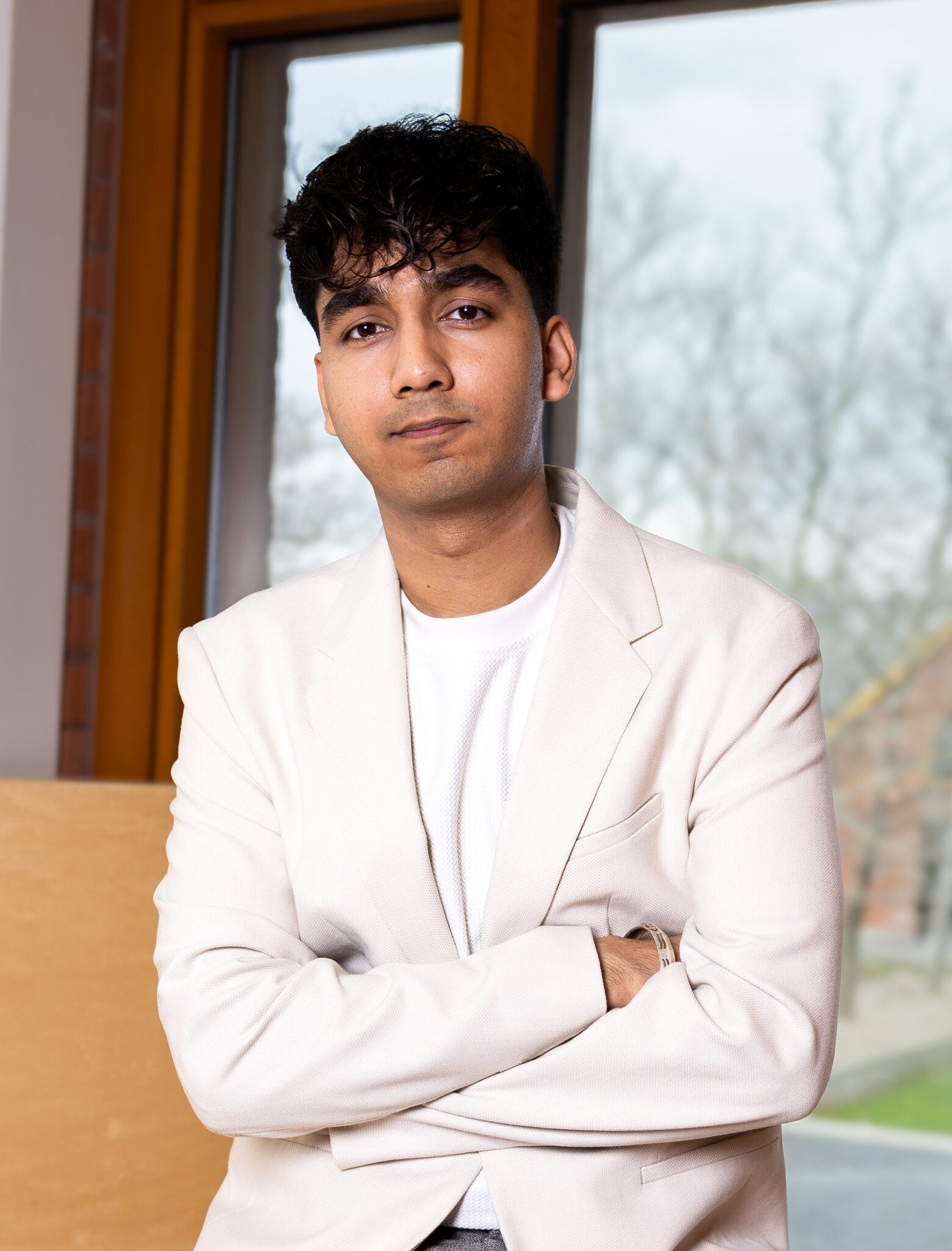A young man with short dark hair, wearing a white blazer and shirt, stands indoors with his arms crossed. A window with a view of trees is visible in the background.