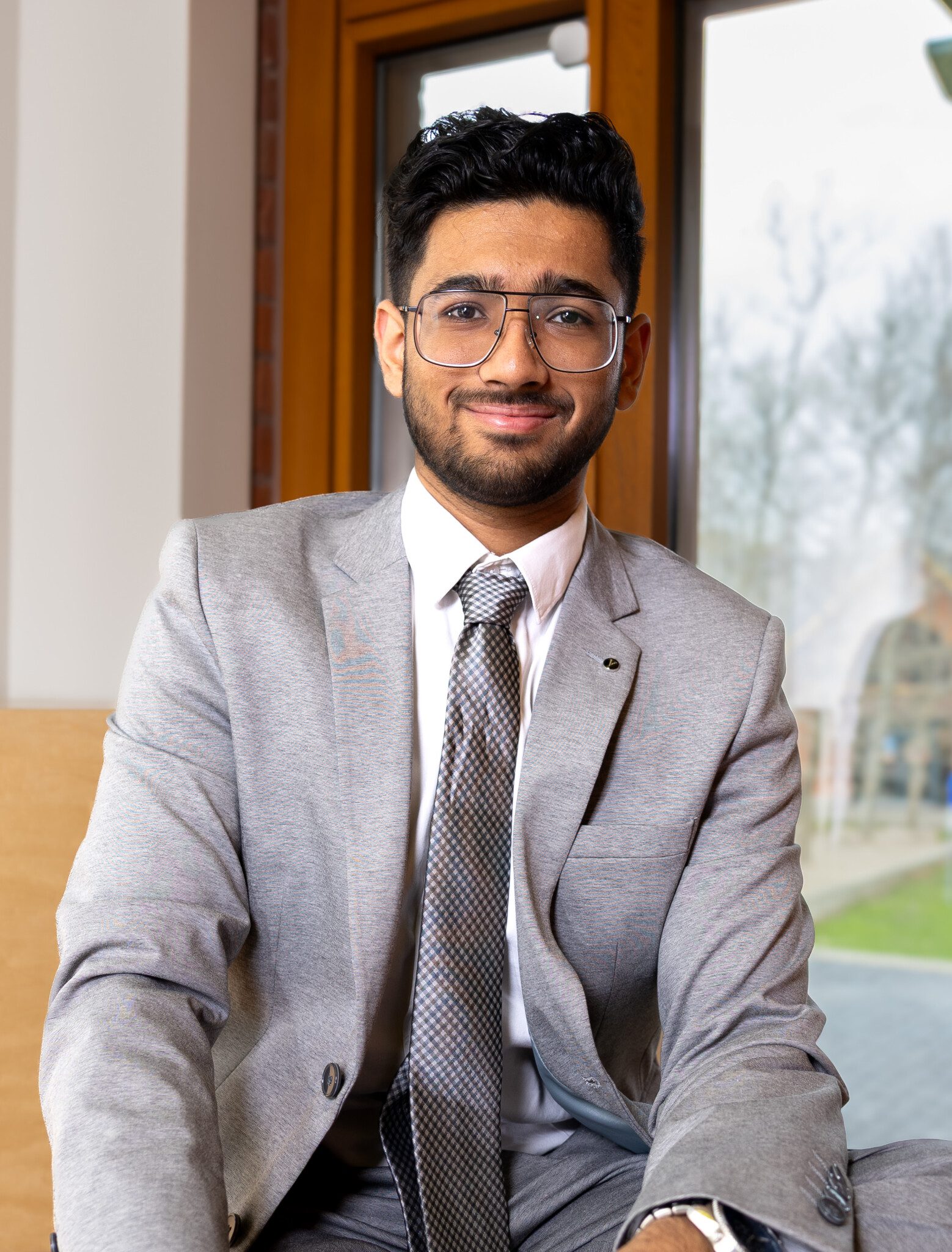 A man wearing a light gray suit, white shirt, and patterned tie sits indoors by a large window, smiling at the camera. He has short black hair, a beard, and is wearing glasses.