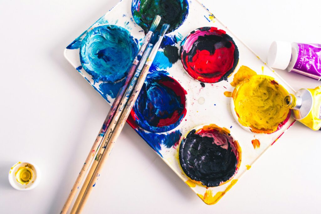 A six cup palette of blue, red, yellow and green paint with three thin messy paint brushes.