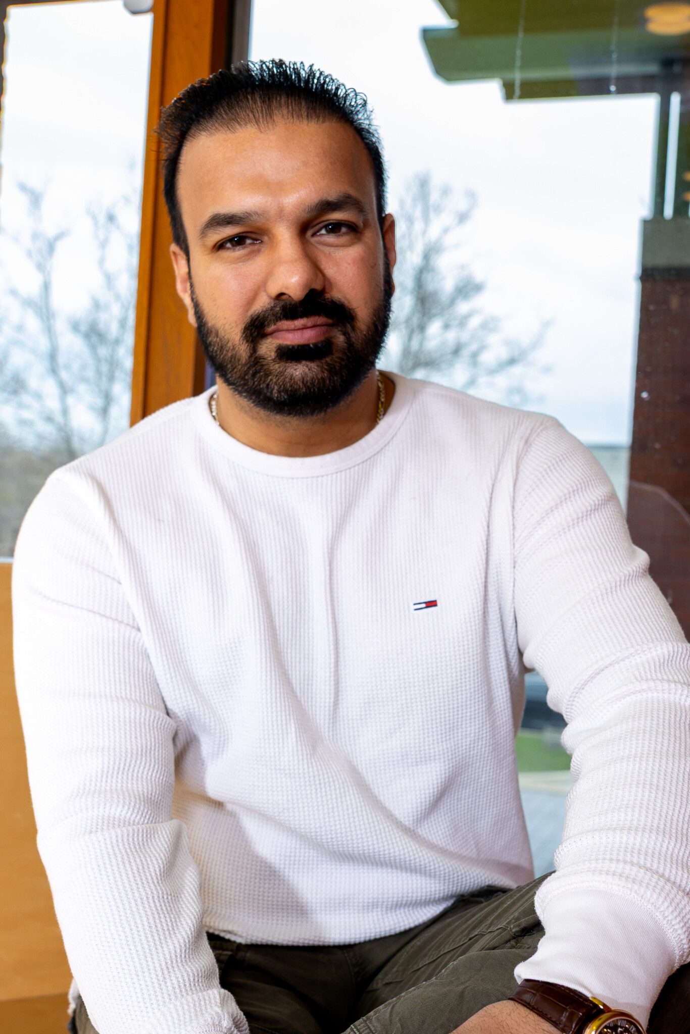 A man with a beard and mustache, wearing a white long-sleeve shirt, sits indoors in front of a window with trees visible outside. He looks at the camera with a relaxed expression.