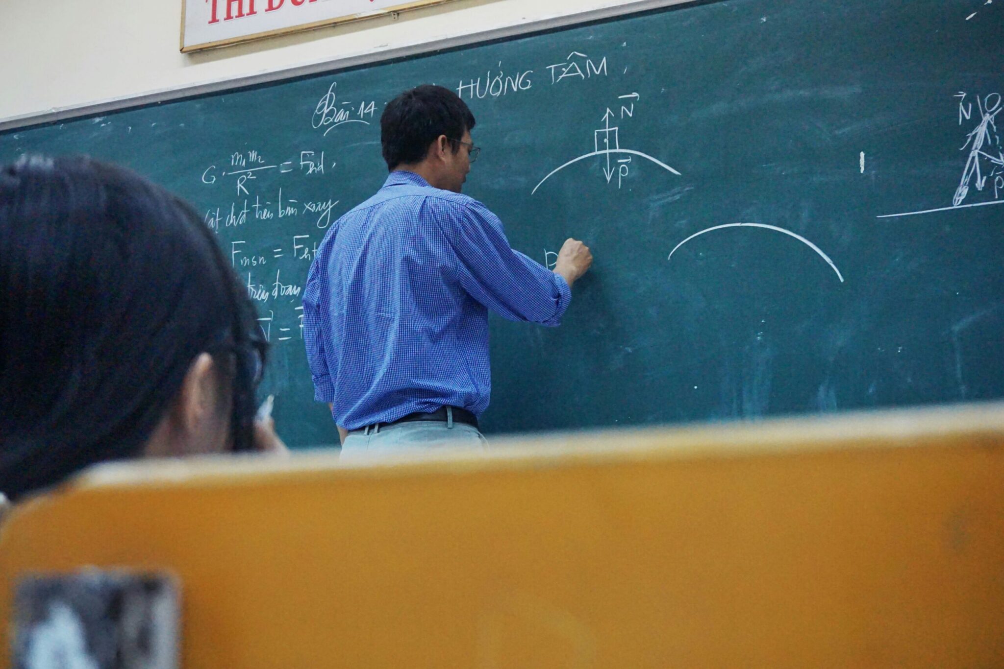An over-the-shoulder shot of a male teacher wearing a blue checkered button-down shirt writing physics equations and drawing force diagrams on a large green chalkboard. In the out-of-focus foreground, a student with dark hair sits at a wooden desk observing the lesson.