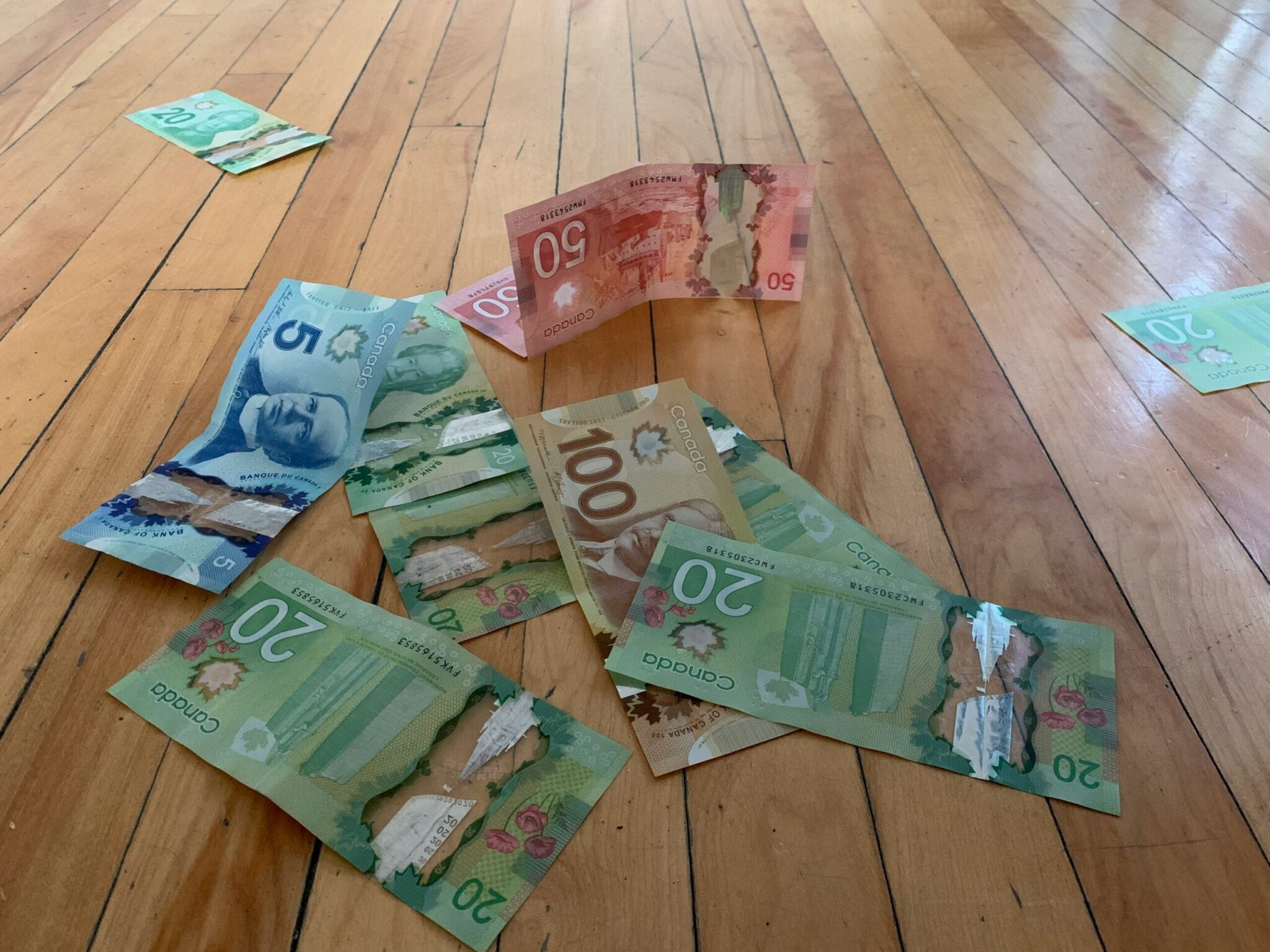 A high-angle shot of various Canadian dollar banknotes scattered across a light-colored hardwood floor. The polymer currency includes several green 20-dollar bills, a blue 5-dollar bill, a brown 100-dollar bill, and a red 50-dollar bill standing upright in the center.