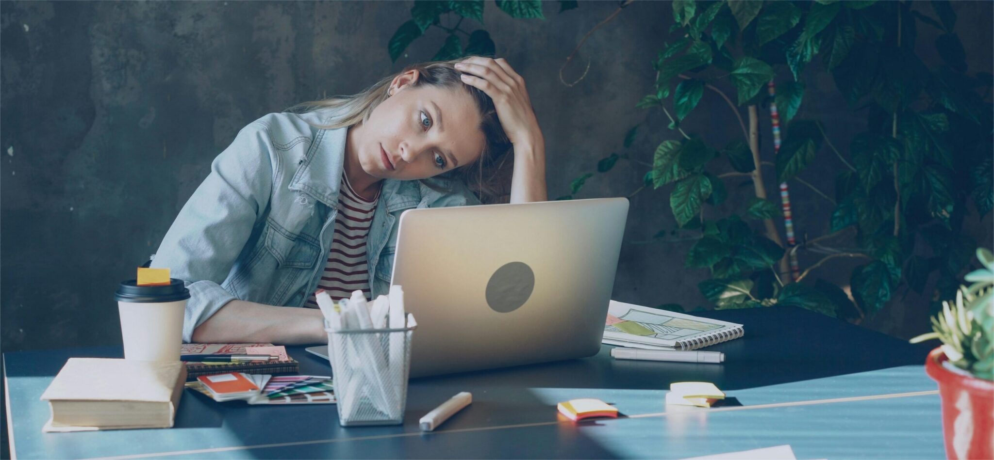 A wide shot of a young blonde woman sitting at a desk and leaning her head on her hand with an overwhelmed expression while staring at her laptop screen. She is wearing a light denim jacket over a striped shirt, surrounded by a coffee cup, stationery, notebooks, color swatches, and sticky notes, with lush green houseplants in the background.