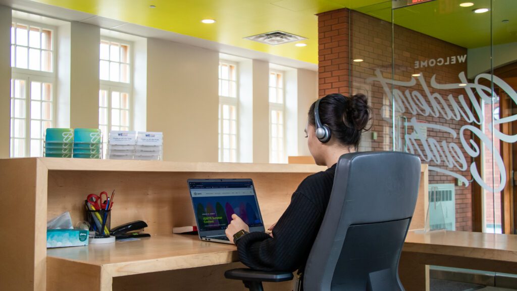A person wearing headphones sits at a desk working on a laptop in a bright, modern office space with large windows, office supplies, and a yellow ceiling.