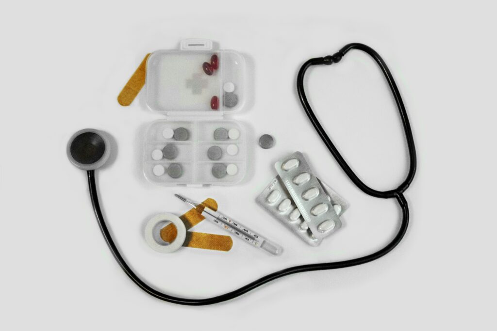 A stethoscope, pillboxes with tablets and capsules, a thermometer, adhesive bandages, medical tape, and blister packs of pills arranged on a white background.