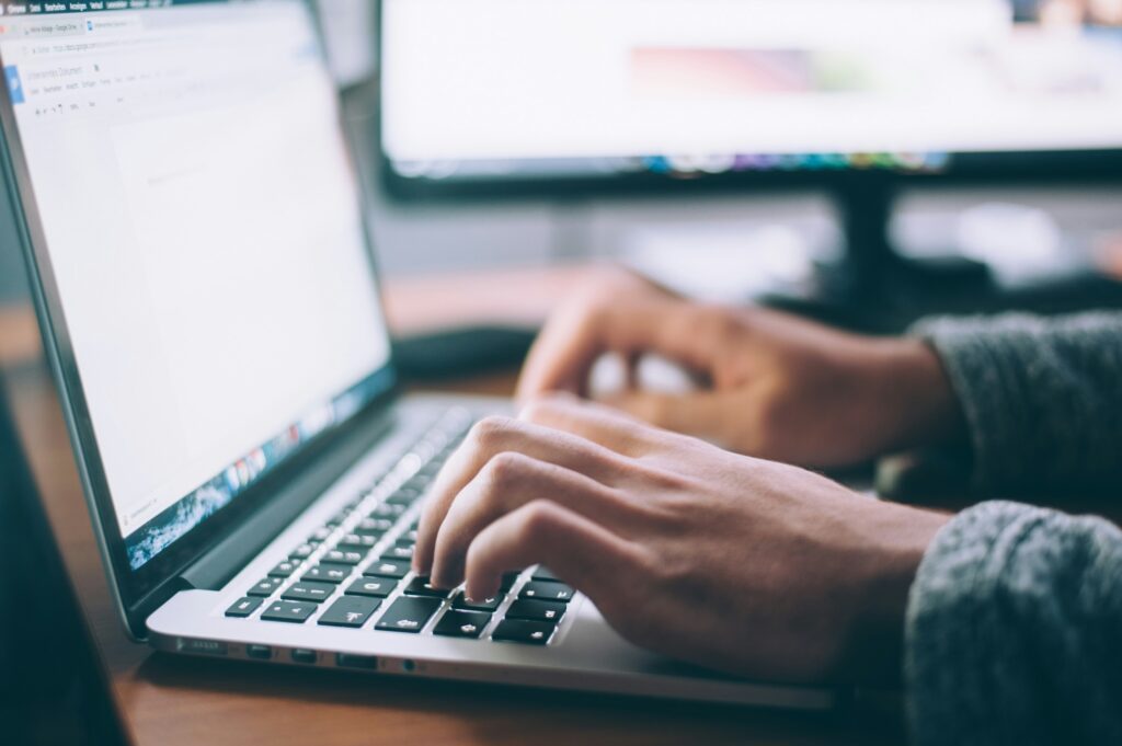 A person typing on a laptop keyboard at a desk, with another computer monitor visible in the background. The focus is on their hands and the laptop.