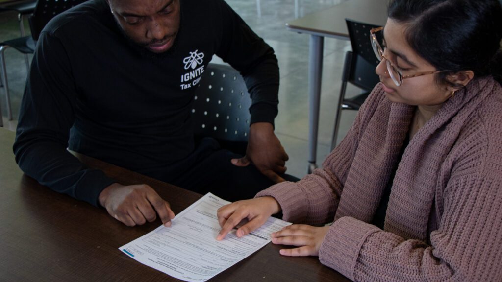 Two people sit at a table, reviewing and discussing a document together. One person points to the paper, while the other looks on attentively, both engaged in the discussion.