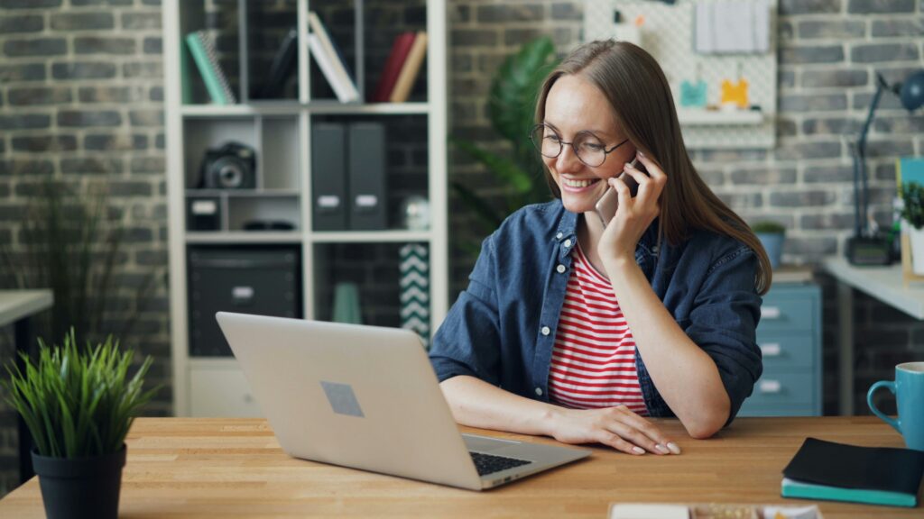 A smiling woman wearing glasses and a striped shirt sits at a desk, talking on the phone while using a laptop. The background shows shelves with office supplies and a plant on the desk.