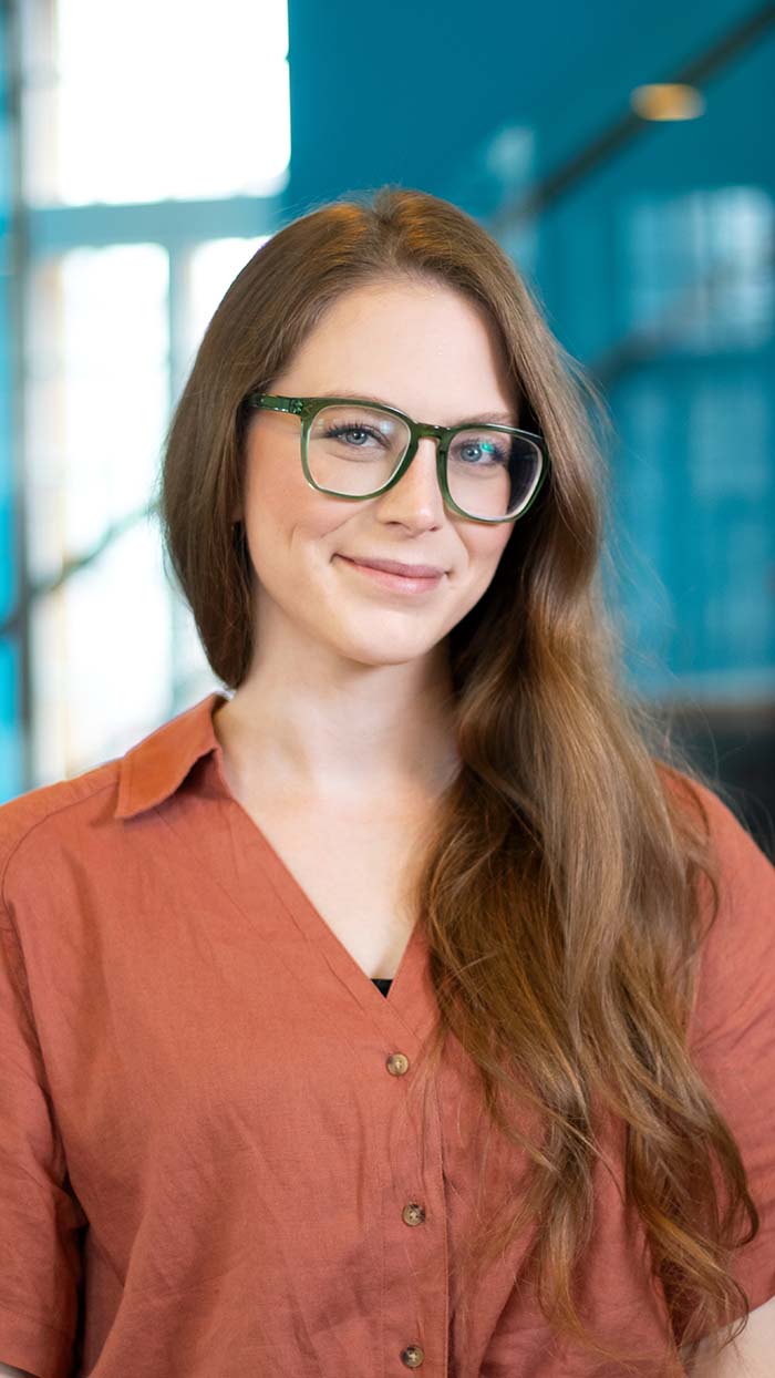 A young woman with long brown hair, wearing green-rimmed glasses and a rust-colored shirt, smiles softly while standing indoors against a blurred blue and windowed background.
