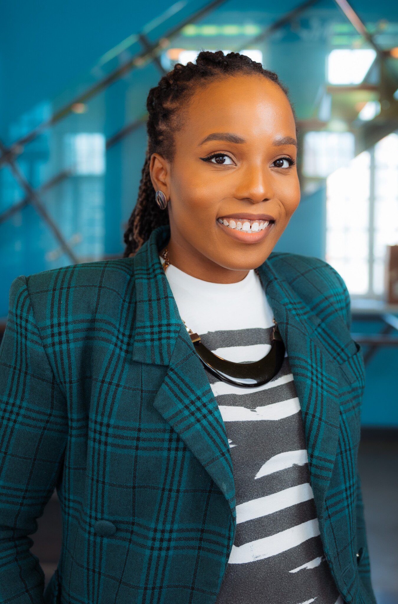 A woman with braided hair smiles, wearing a green plaid blazer, a patterned shirt, and a black necklace, standing indoors with a blurred blue and windowed background.