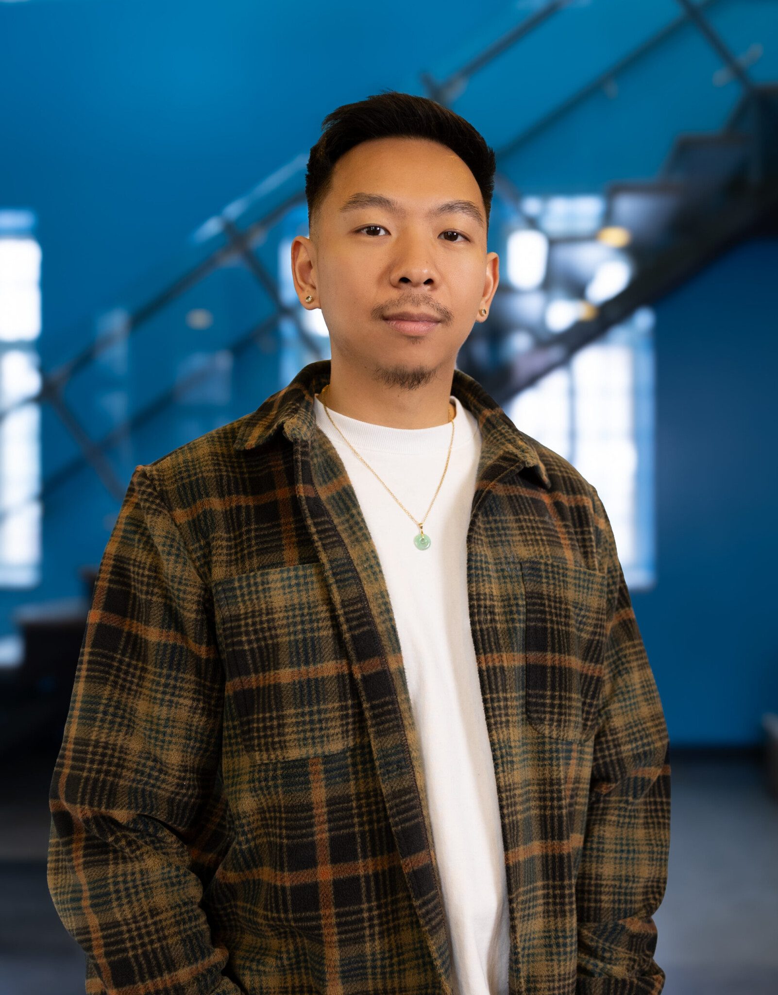 A man with short black hair and facial hair wears a plaid shirt over a white t-shirt, standing indoors in front of a modern blue wall and staircase, looking confidently at the camera.