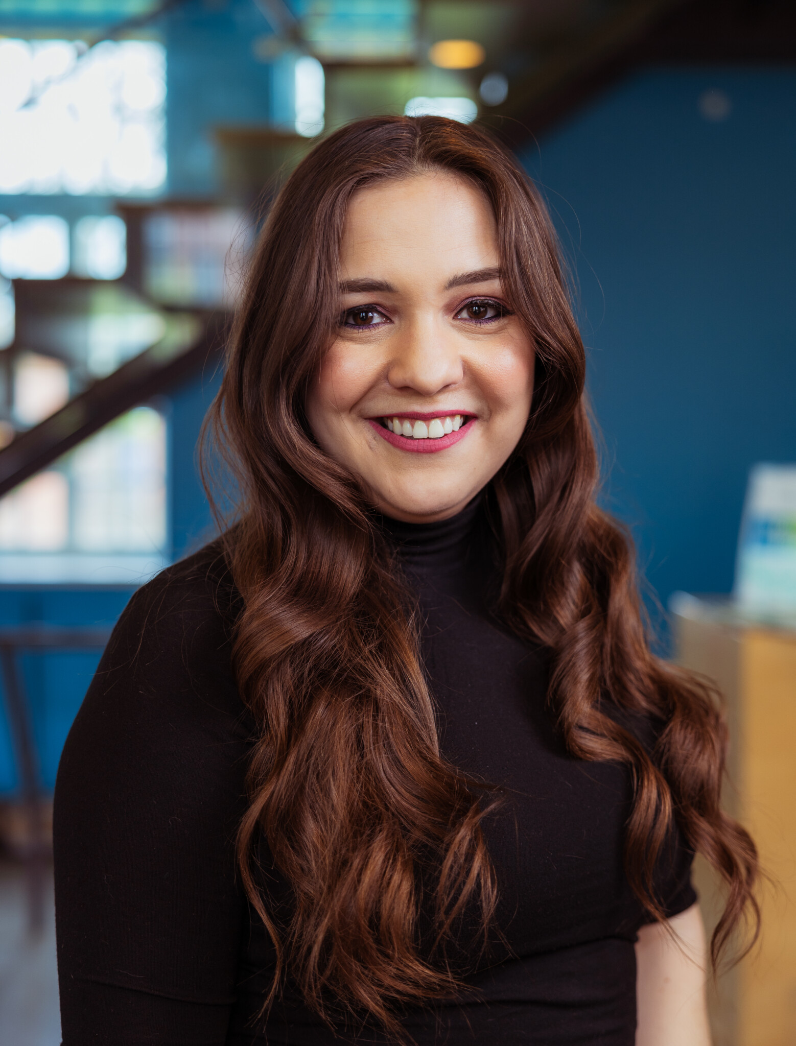 A woman with long wavy brown hair, wearing a black top, smiles at the camera in an indoor setting with blue walls and blurred background details.