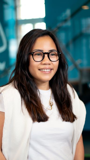 A woman with long dark hair and glasses smiles at the camera. She is wearing a white shirt, a light-colored sweater draped over her shoulders, and a gold necklace. The background is blue and slightly blurred.