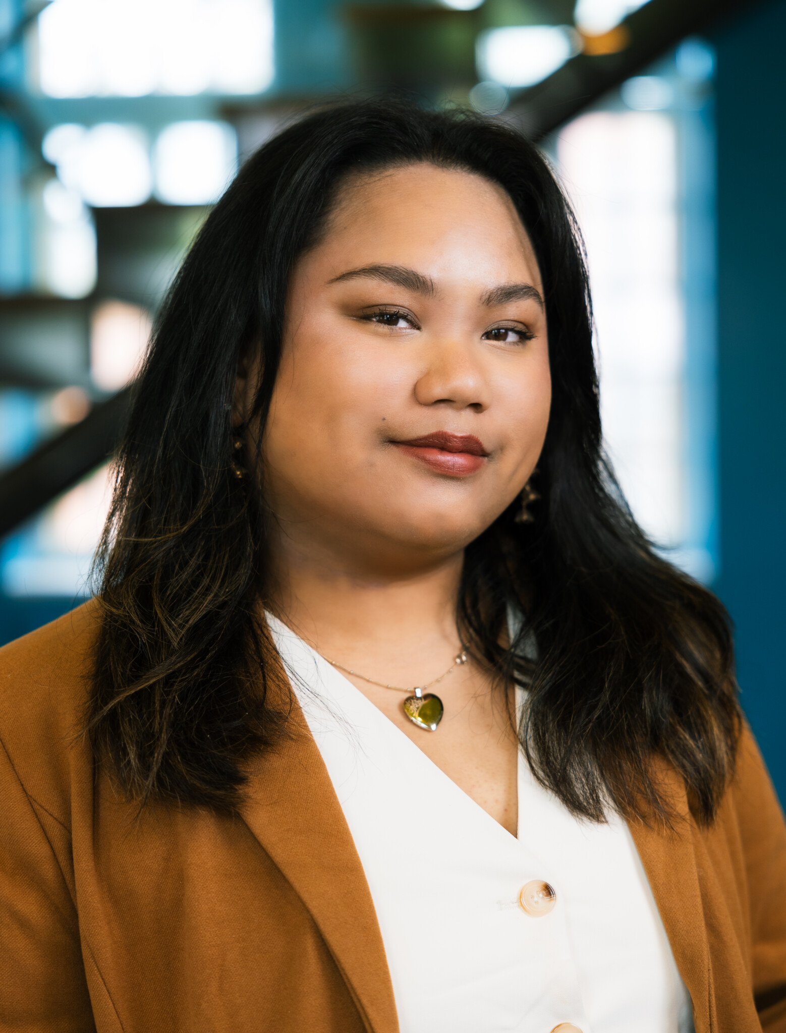 A woman with long dark hair wearing a brown blazer and white top stands indoors, looking confidently at the camera. The background is softly blurred with blue and natural light.