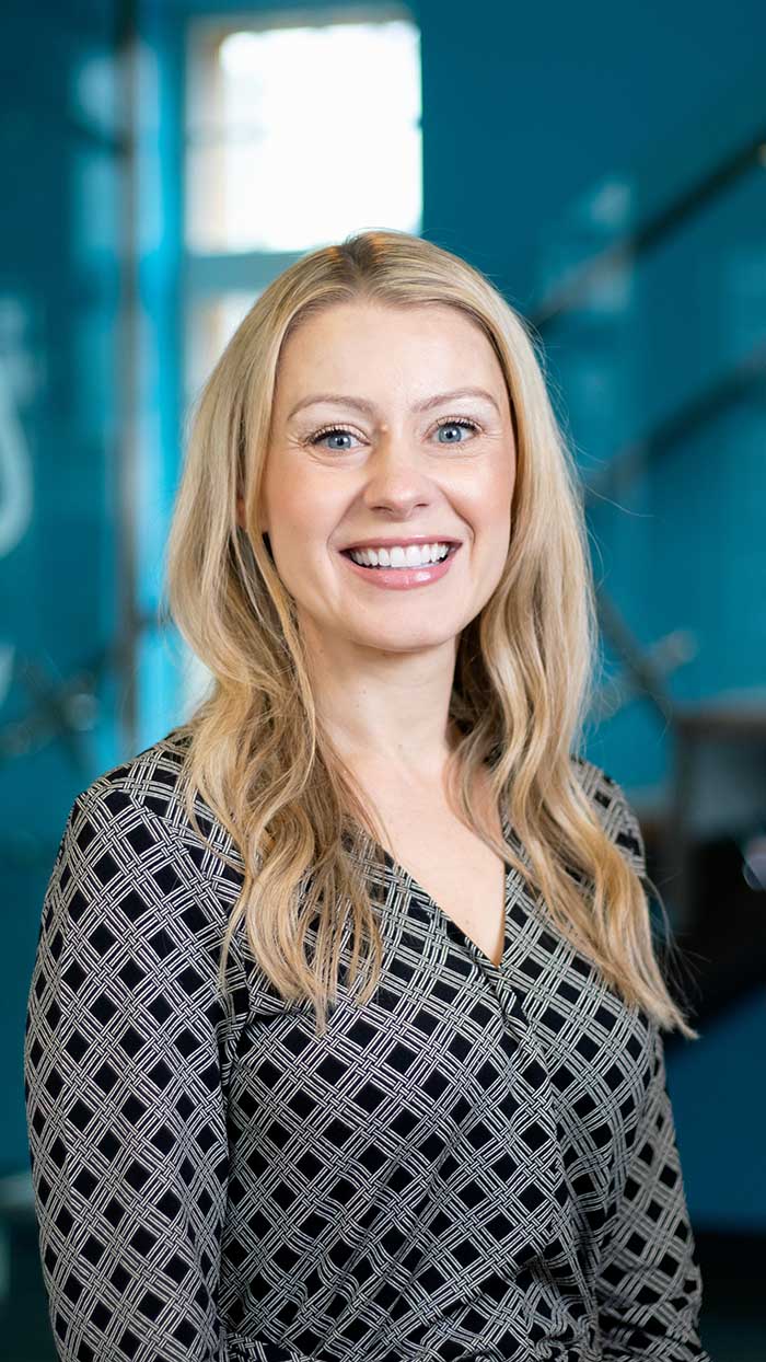 A woman with long blonde hair wearing a black and white patterned blouse smiles at the camera in a brightly lit indoor setting with a blue background.